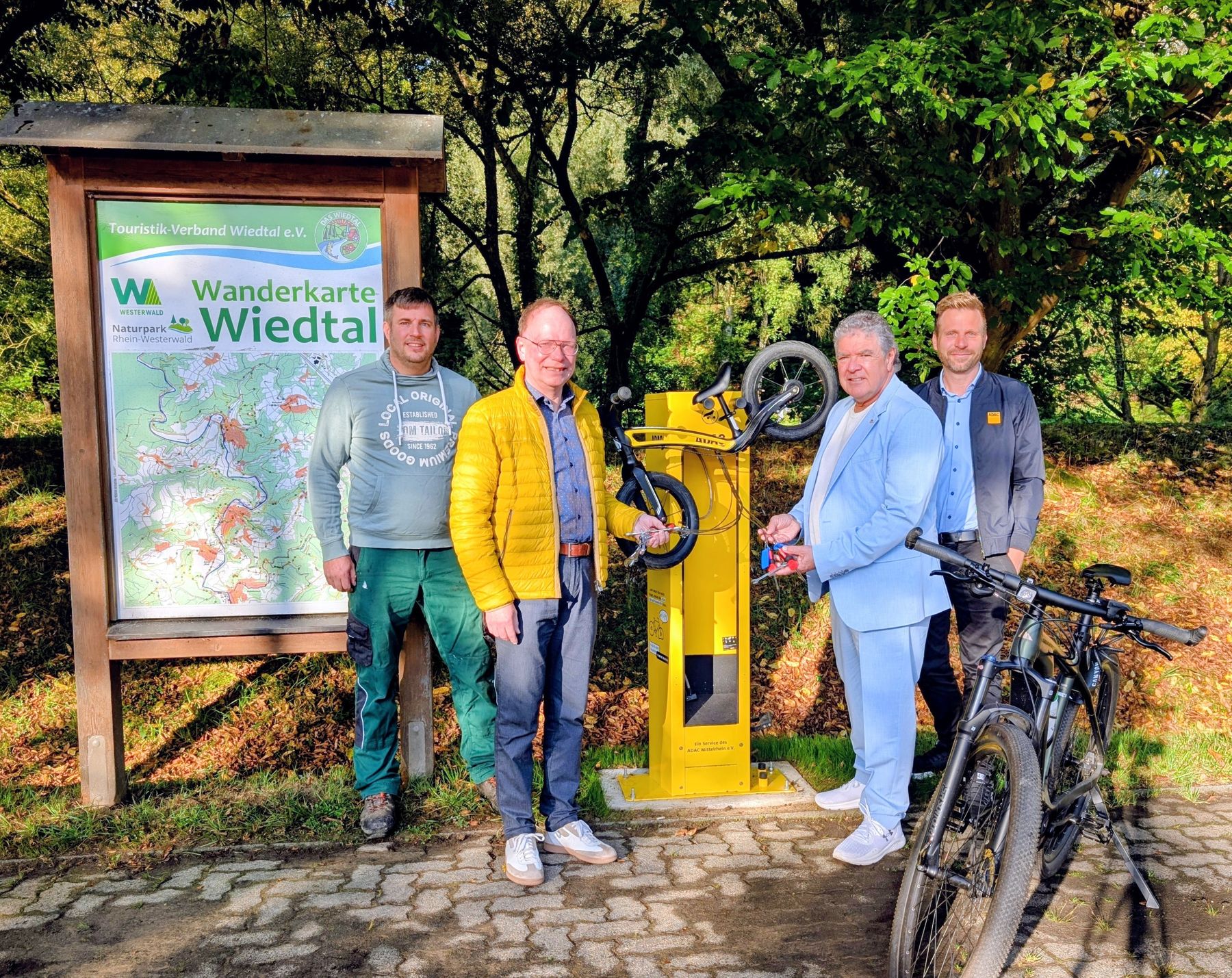 Four men stand beside a yellow bicycle repair station and two bicycles, in front of a large map sign titled Wanderkarte Wiedtal, surrounded by lush green trees on a sunny day.